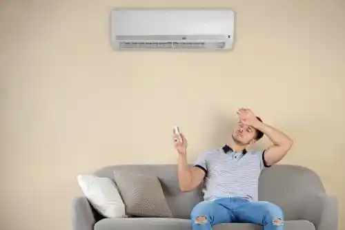 A view of a guy sitting under an ac unit holding a remote wiping his sweat